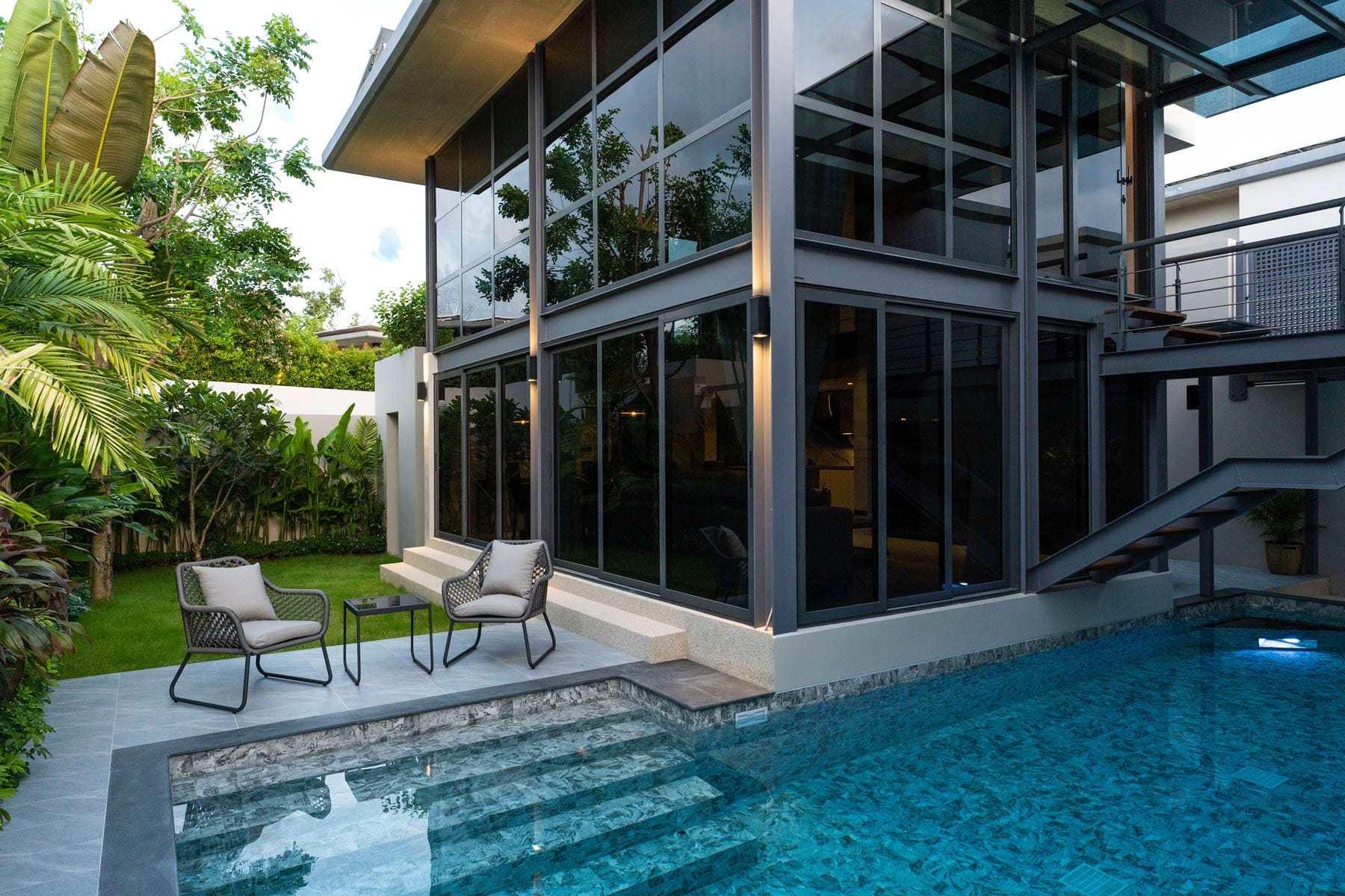 Modern glass-front villa with private pool, outdoor lounge area, and tropical garden landscaping.