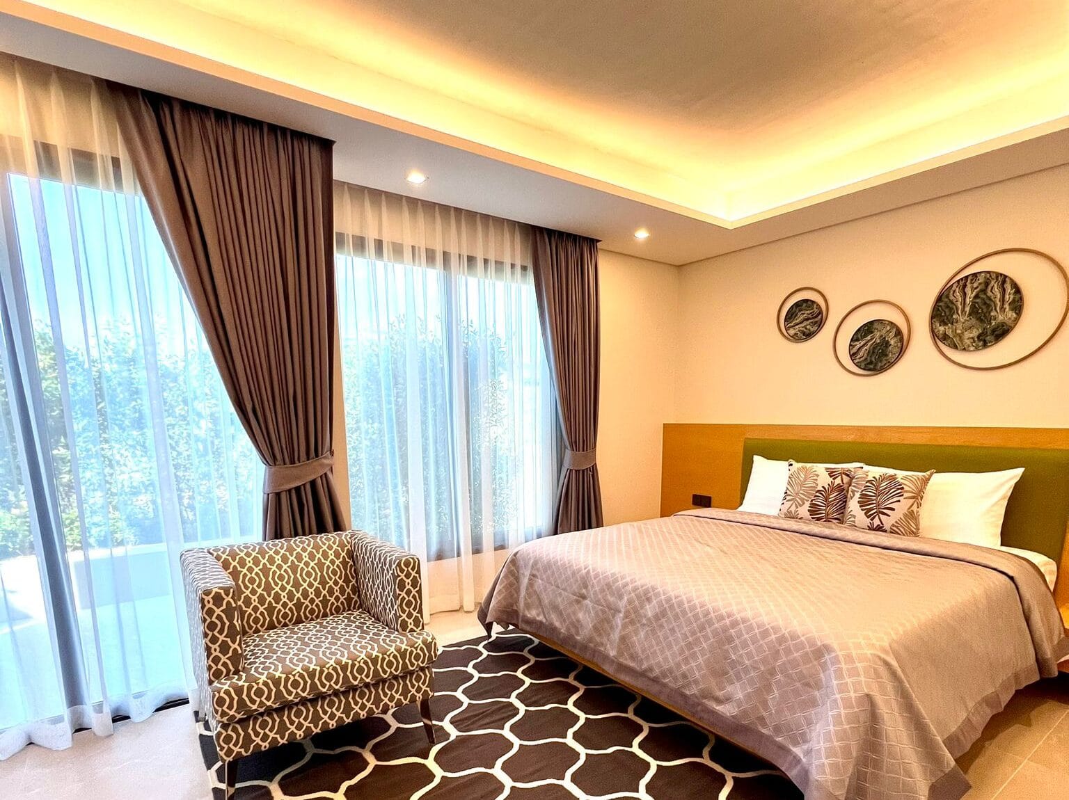 Modern bedroom with king-size bed, patterned armchair, large windows with curtains, and soft ambient lighting in a villa at RIVERHOUSE Phuket.